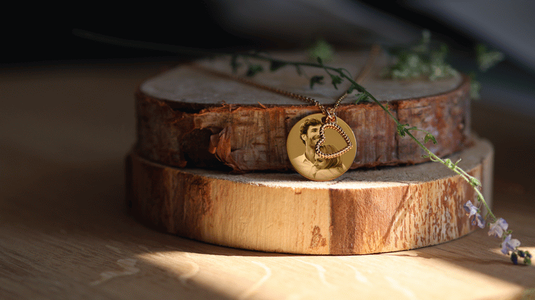 Pendentif gravé en plaqué or sur un support en bois, entouré de plantes délicates, symbolisant l'élégance et la personnalisation.