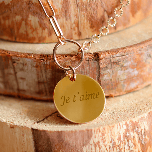 Collier plaqué or rond gravé avec 'Je t'aime' sur un fond en bois. Parfait pour un cadeau personnalisé.