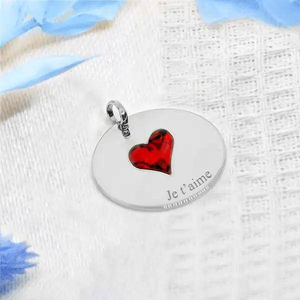 Petit médaillon rond en argent avec cœur rouge gravé "Je t'aime" sur fond textile clair.