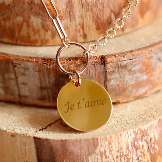 Collier plaqué or rond gravé avec 'Je t'aime' sur un fond en bois. Parfait pour un cadeau personnalisé.