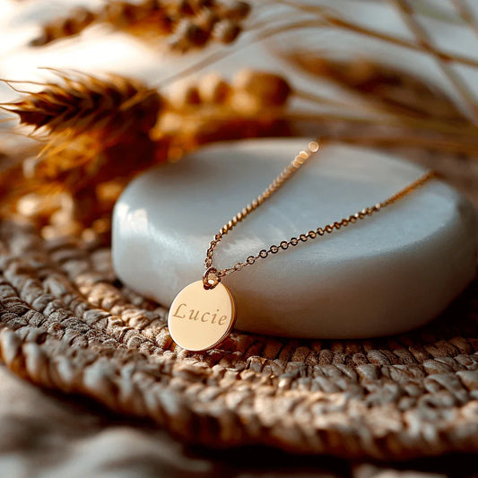 Médaille en acier doré gravée avec le prénom Lucie, posée sur une pierre douce, entourée de décoration naturelle.
