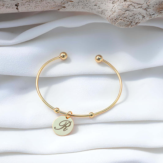 Bracelet jonc doré GravurePhoto avec pendentif rond personnalisé lettre R, bijou élégant sur tissu blanc.